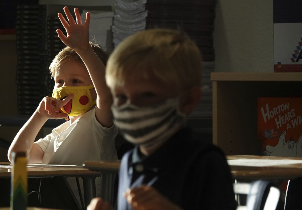 A masked first-grader raises his hand in class.