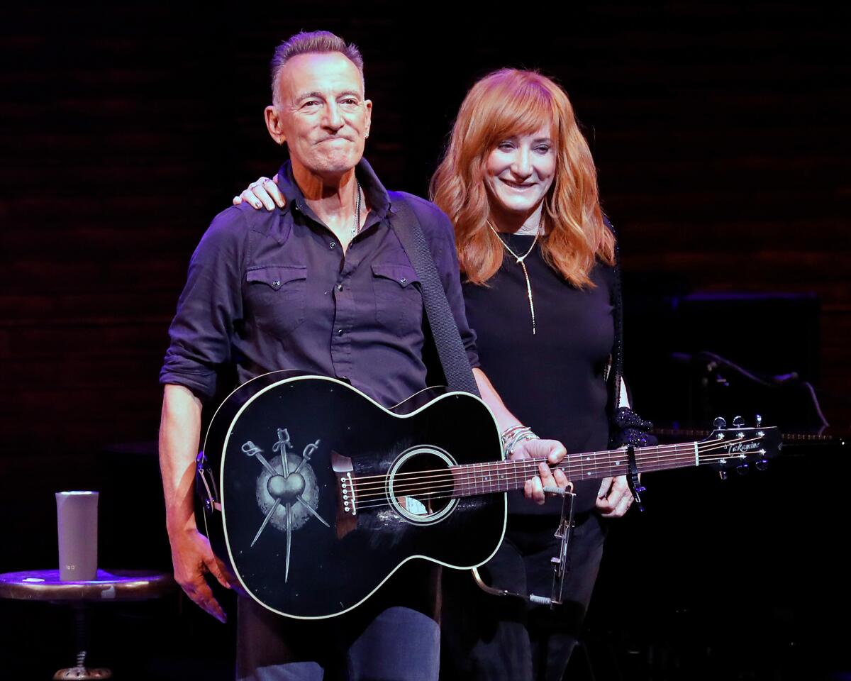 A man holding a guitar and a woman with her arm around him stand on a stage