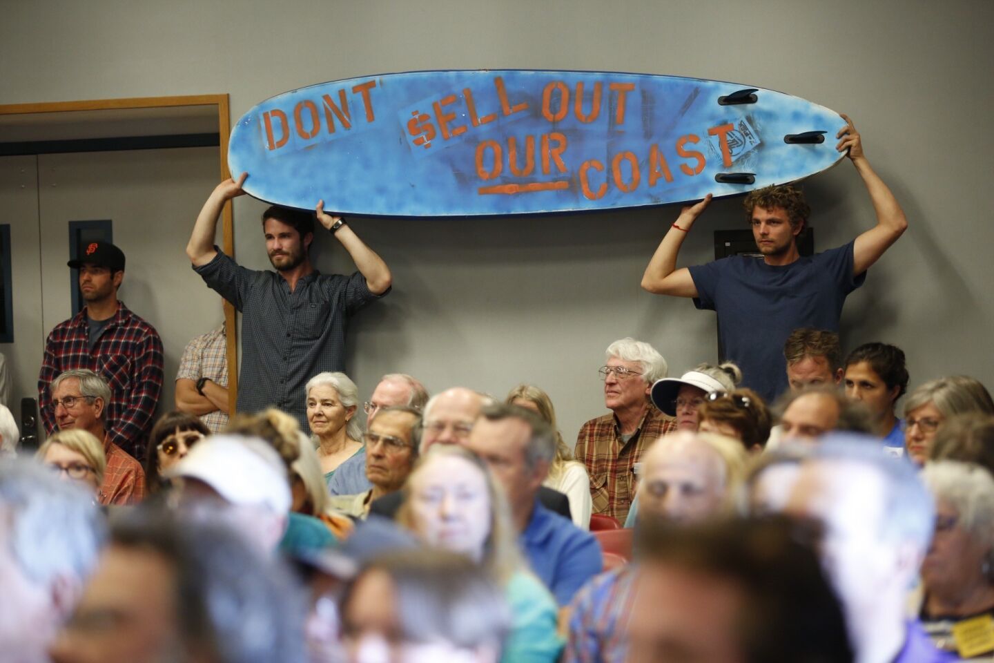 Alex Lowe, left, and Rob Moddlemog from Orange County hold a surf board as members of the California Coastal Commission listen to speakers in support of Charles Lester.