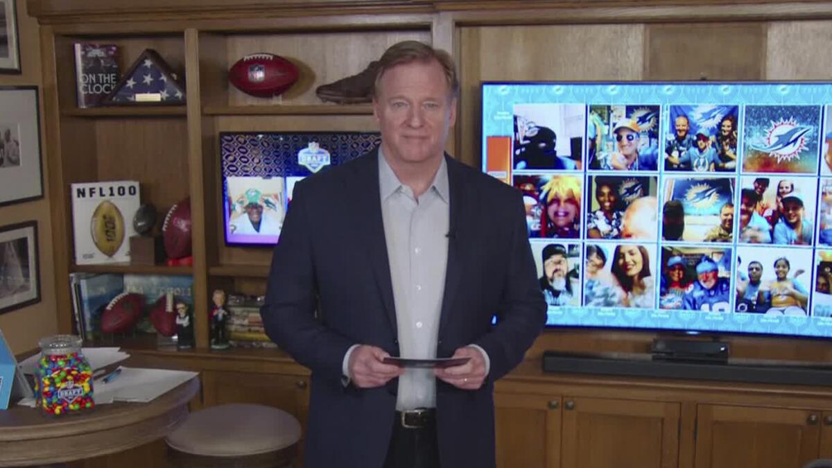 Roger Goodell stands in a room with footballs and a big TV screen