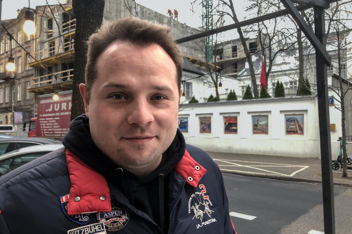 A smiling man with dark brown hair and wearing a dark jacket stands in front of buildings