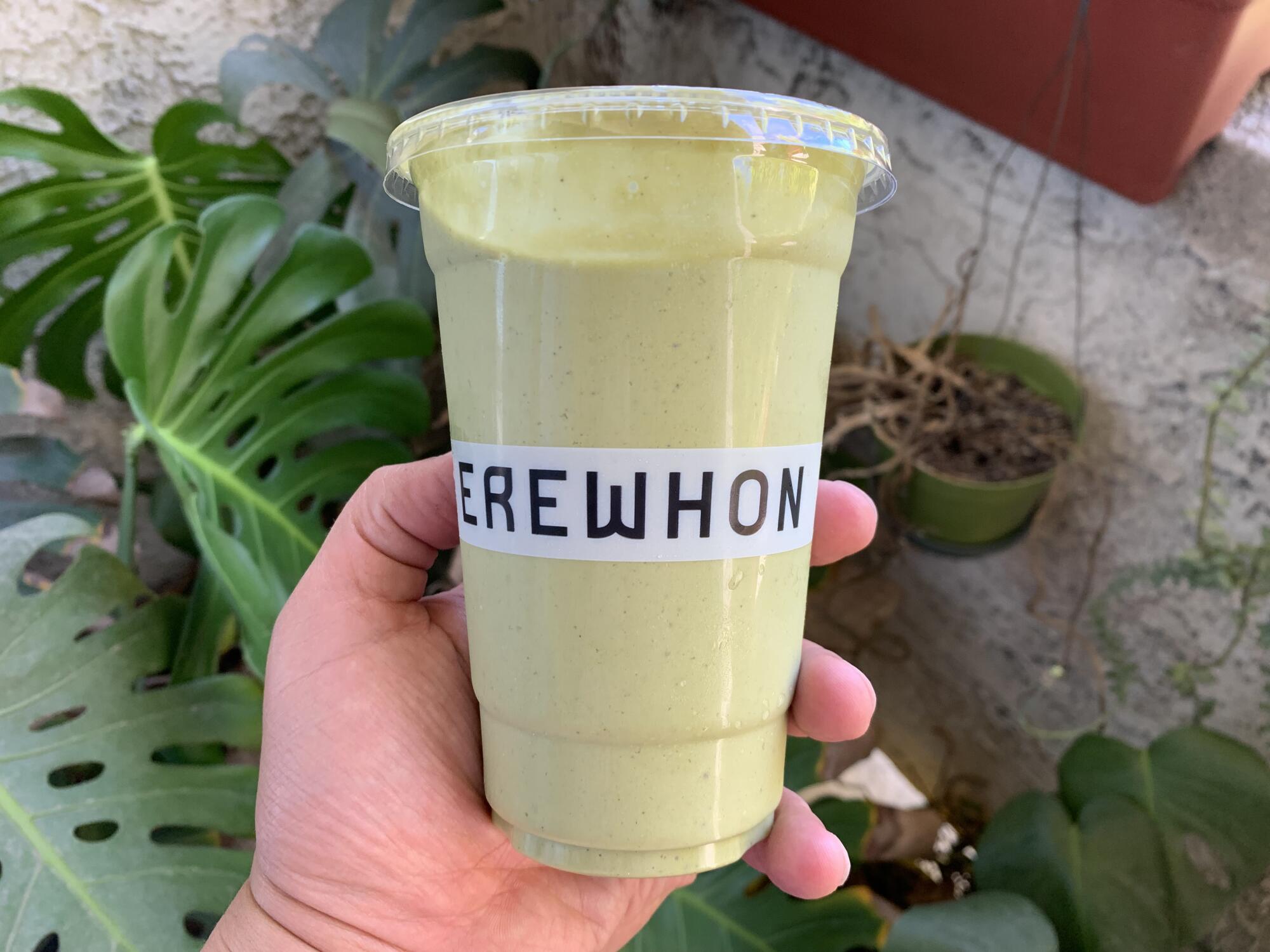 A hand holds a yellowish-green smoothie in a plastic cup that says Erewhon.