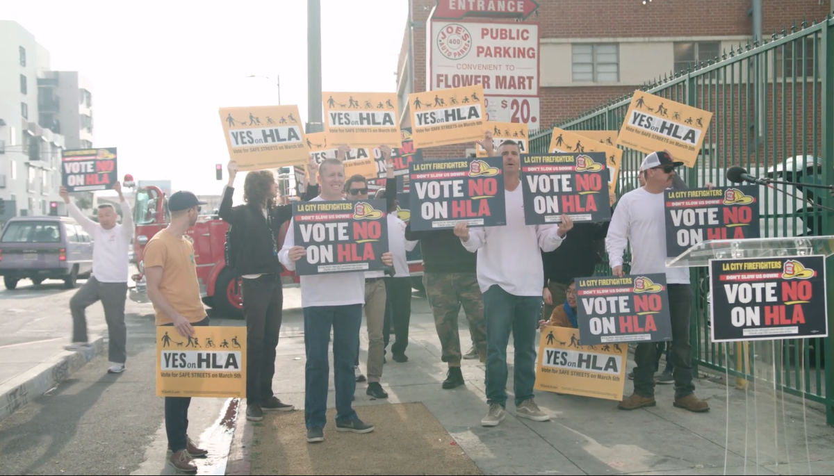 People stand on a sidewalk holding signs for and against ballot measure HLA.