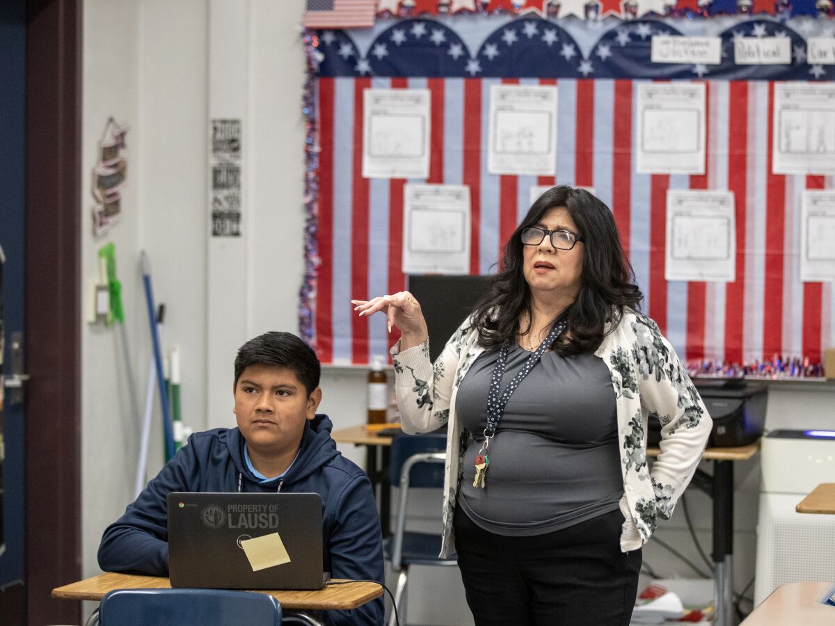 A male student, seated, next to a female teacher, standing.