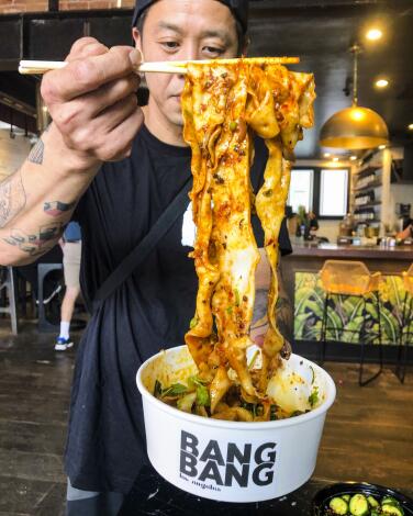 A man uses chopsticks to hold up broad, saucy noodles from a container that says Bang Bang