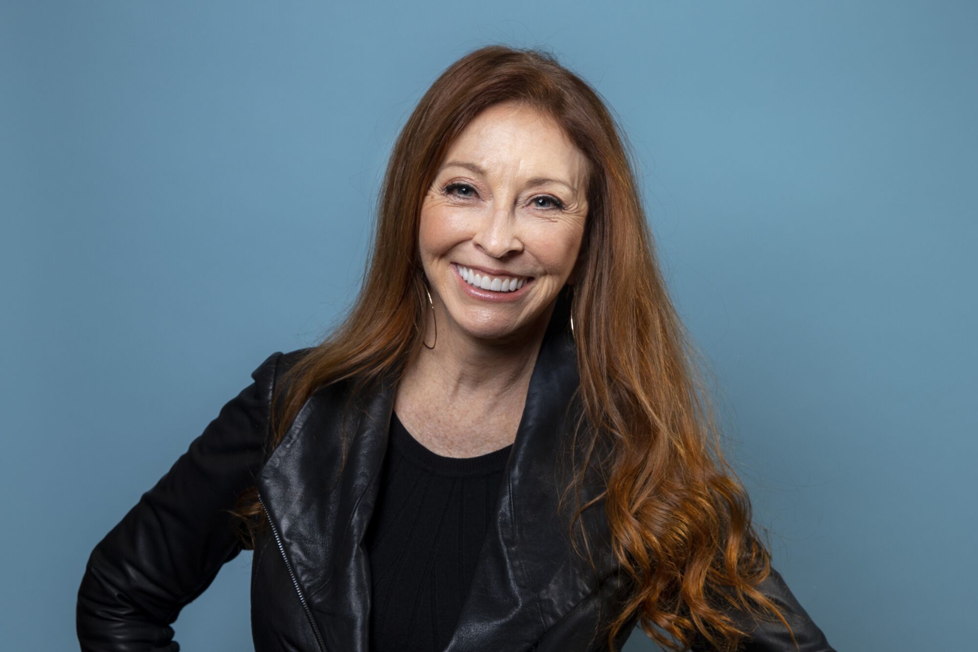 Cassandra Peterson, wearing all black with long red hair, smiles with her hands on her hips