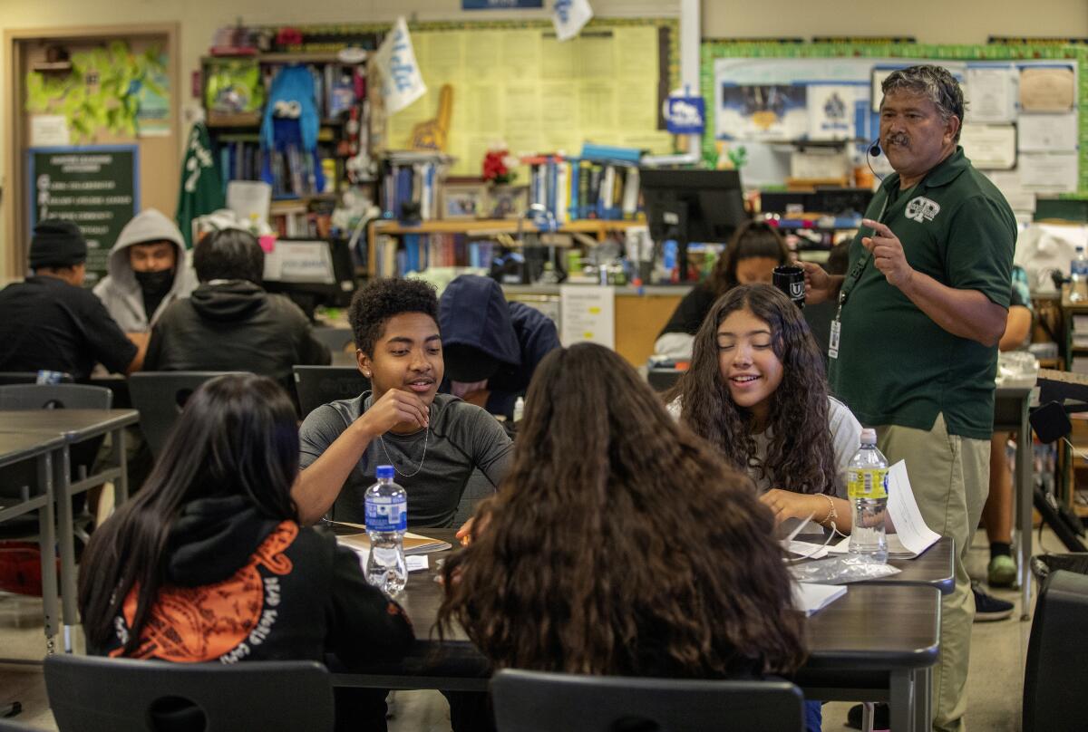 A man in a green short-sleeve shirt and khaki pants stands near four students seated at desks in a classroom