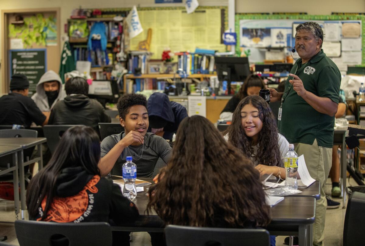 A man in a green short-sleeve shirt and khaki pants stands near four students seated at desks in a classroom 