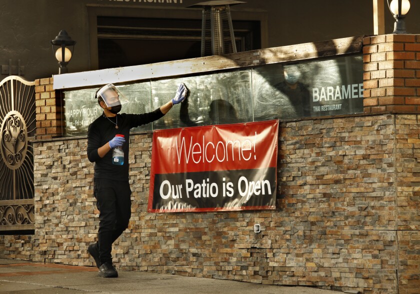 A man in a mask and face shield wipes glass above a banner that says "Welcome, our patio is open"