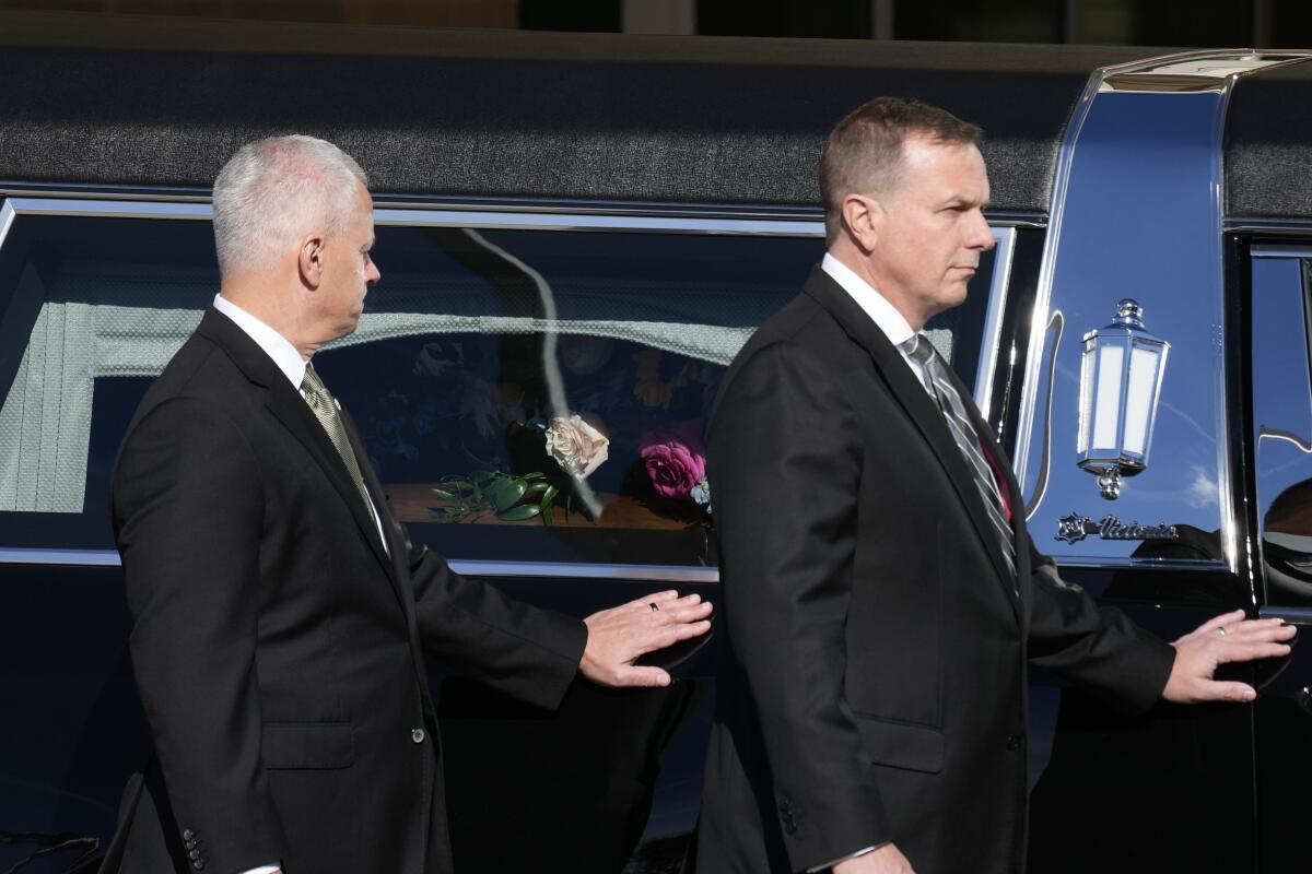 Men in black suits stand with their hand on a hearse 