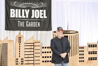 Billy Joel in a black shirt and blazer, jeans and a hat posing infant of a cloth backdrop