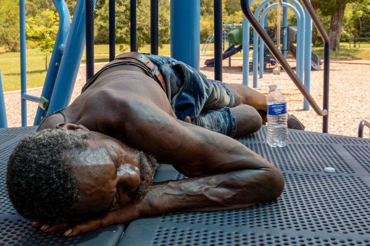 A shirtless man with a bottle of water, lying in the shade on a playground set's raised platform.