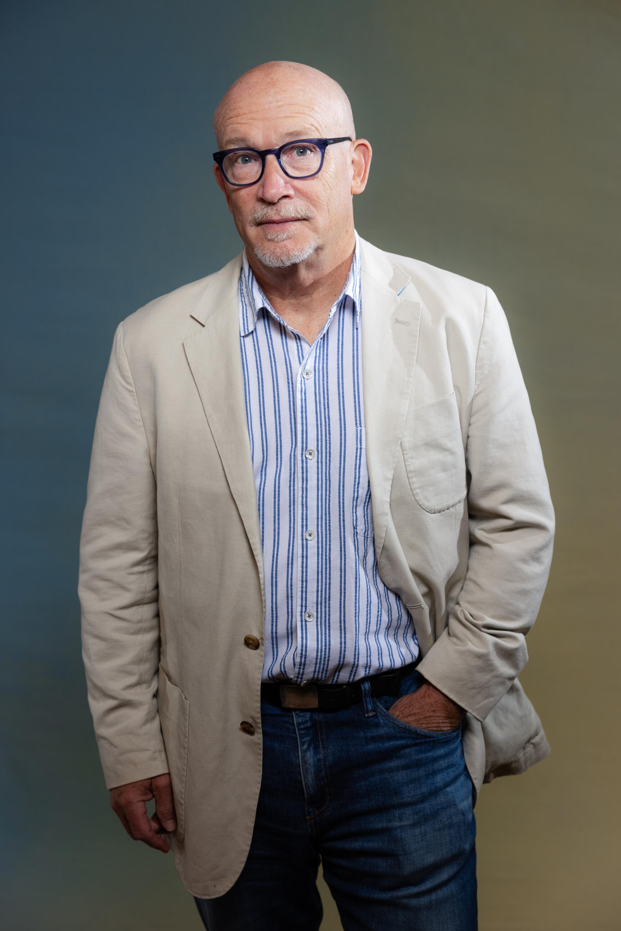 Alex Gibney stands with a hand in his pants pocket while wearing a striped shirt and jacket.