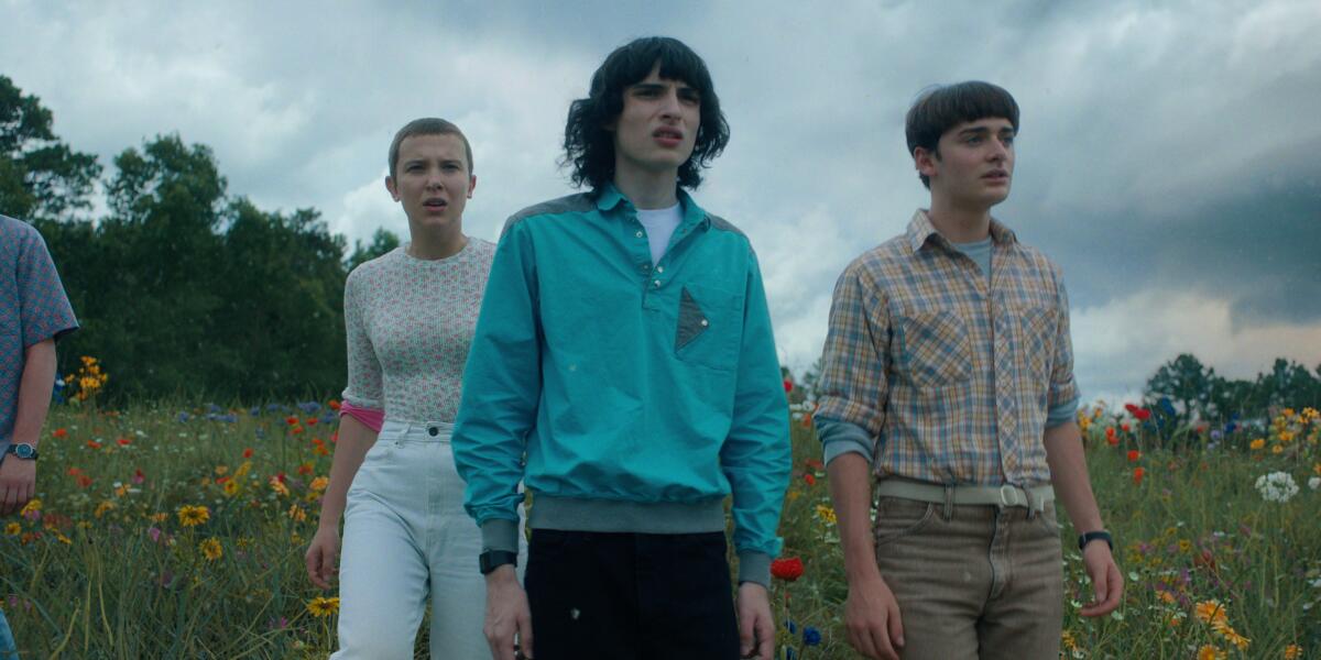 Millie Bobby Brown, Finn Wolfhard and Noah Schnapp walk through a flowery field in their 'Stranger Things' costumes.