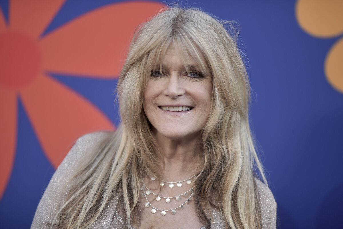A smiling woman with long straight blond hair, wearing a beige outfit and silver jewelry.