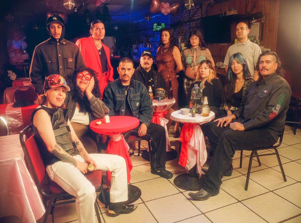 A group of Latinx artists seated at or standing behind bar tables draped in red fabric at a small downtown L.A. bar.