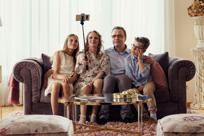 A family of four sits on a couch, with the mother holding up a selfie stick holding a phone 