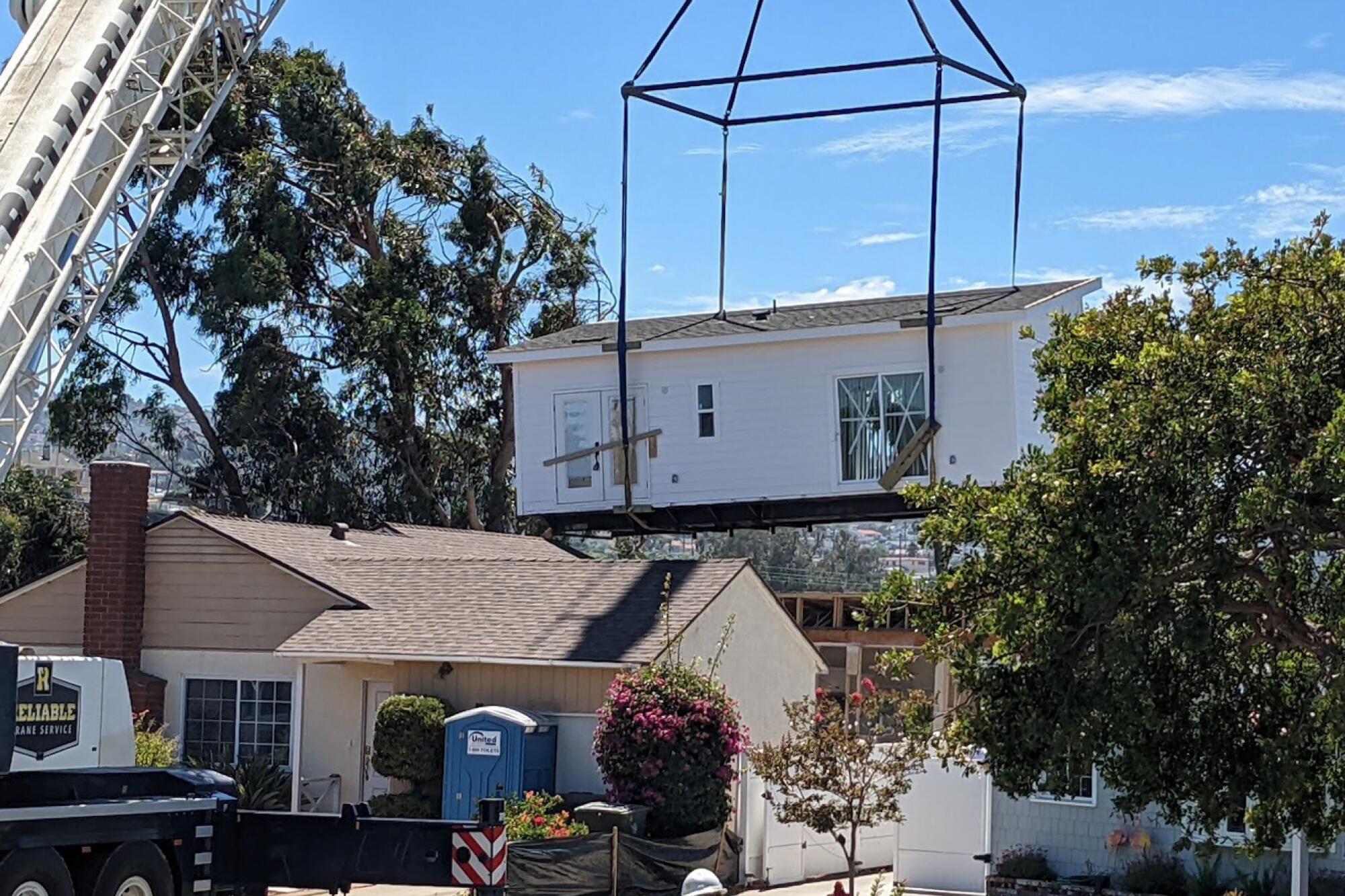 One half of a prefab building is moved into position behind a house with the help of a crane.