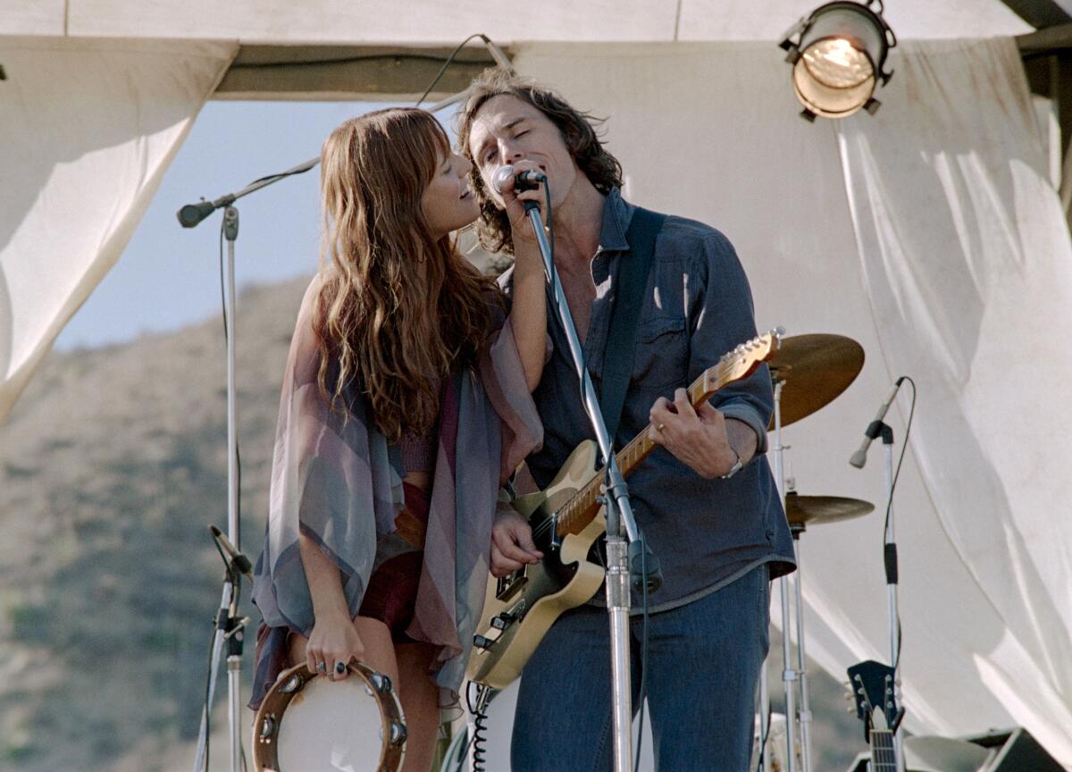 A woman with long hair and holding a tambourine and a man playing a guitar sing into a microphone on an outdoor stage.