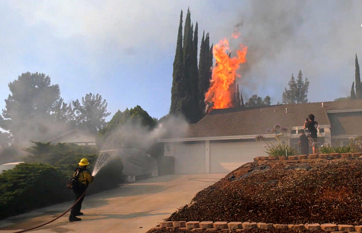 A firefighter standing on a driveway aims a hose at a house with flames seen behind it 