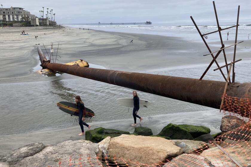 Surfers walk under a pipeline installed to carry sand from the harbor to beaches near the pier.