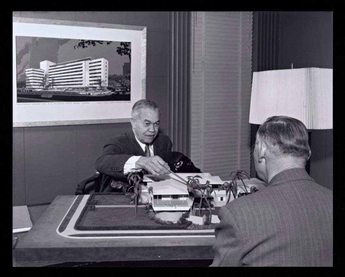 In a black-and-white image, architect Paul Williams is seen pointing at a model while seated at his desk