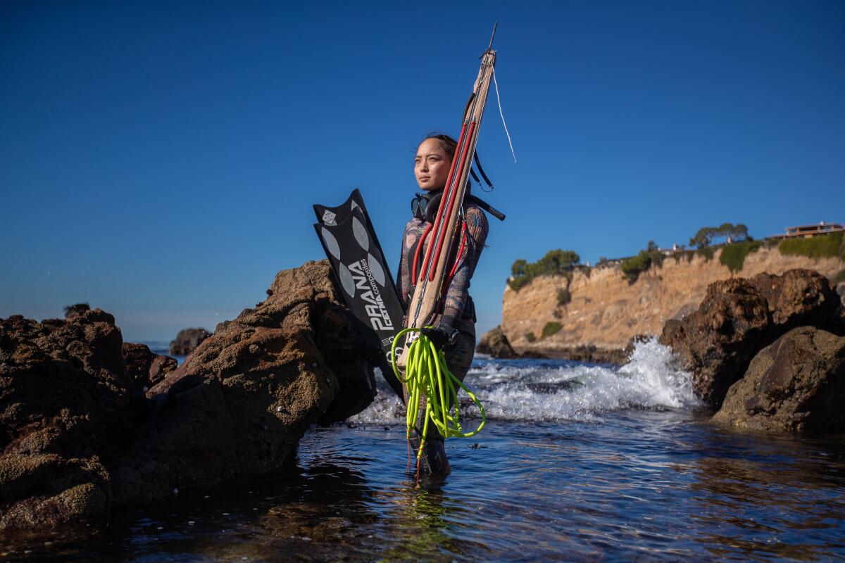 A woman in a wetsuit with a spear stands in the ocean among rocks