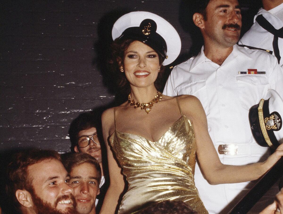 A woman in a tight-fitting gold gown wearing a sailor's hat surrounded by men in uniform.