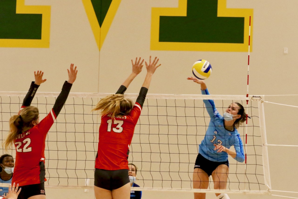 A female volleyball player jumps to spike a ball and defensive players jump to block.