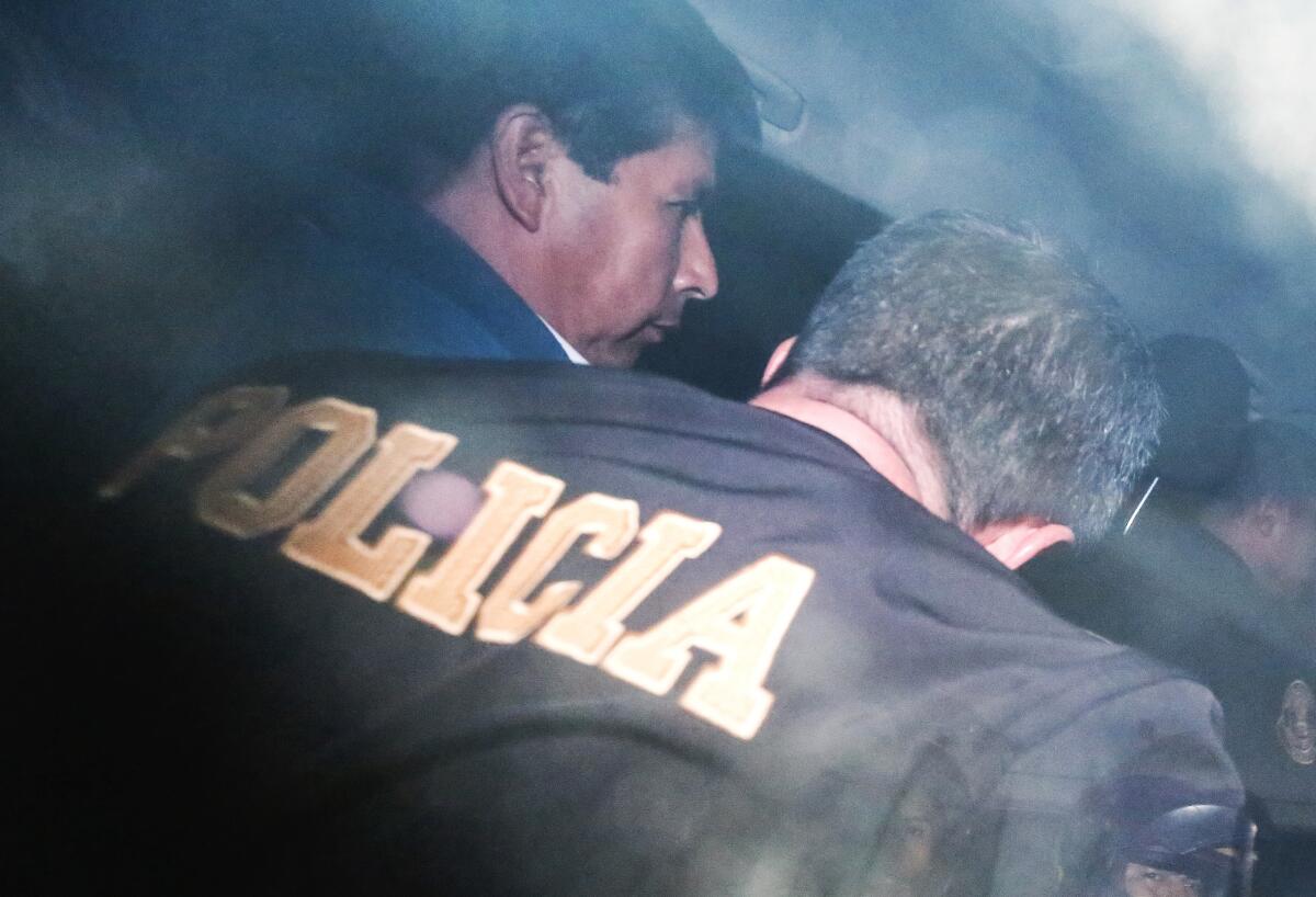 A view from behind of a man with dark hair escorted by a man in a jacket with the word Policia 