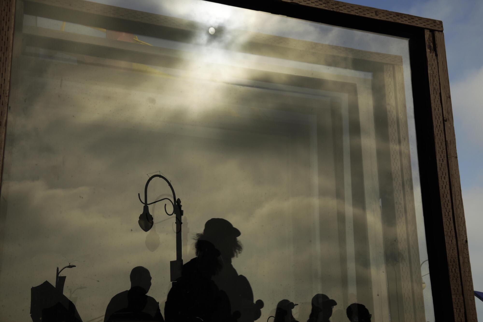 People, reflected on glass panels, make their way through the muggy weather on the Santa Monica Pier 