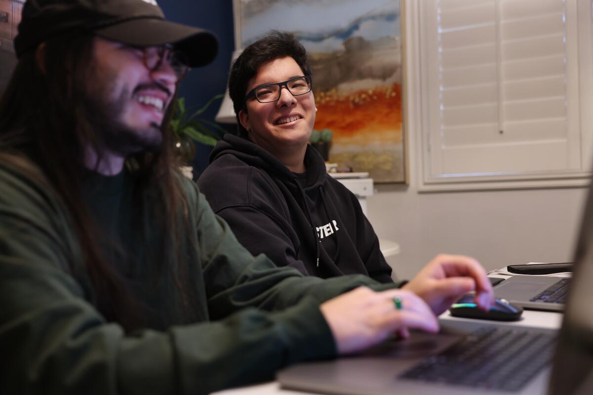 A man wearing a hat and another man smile while working on computers