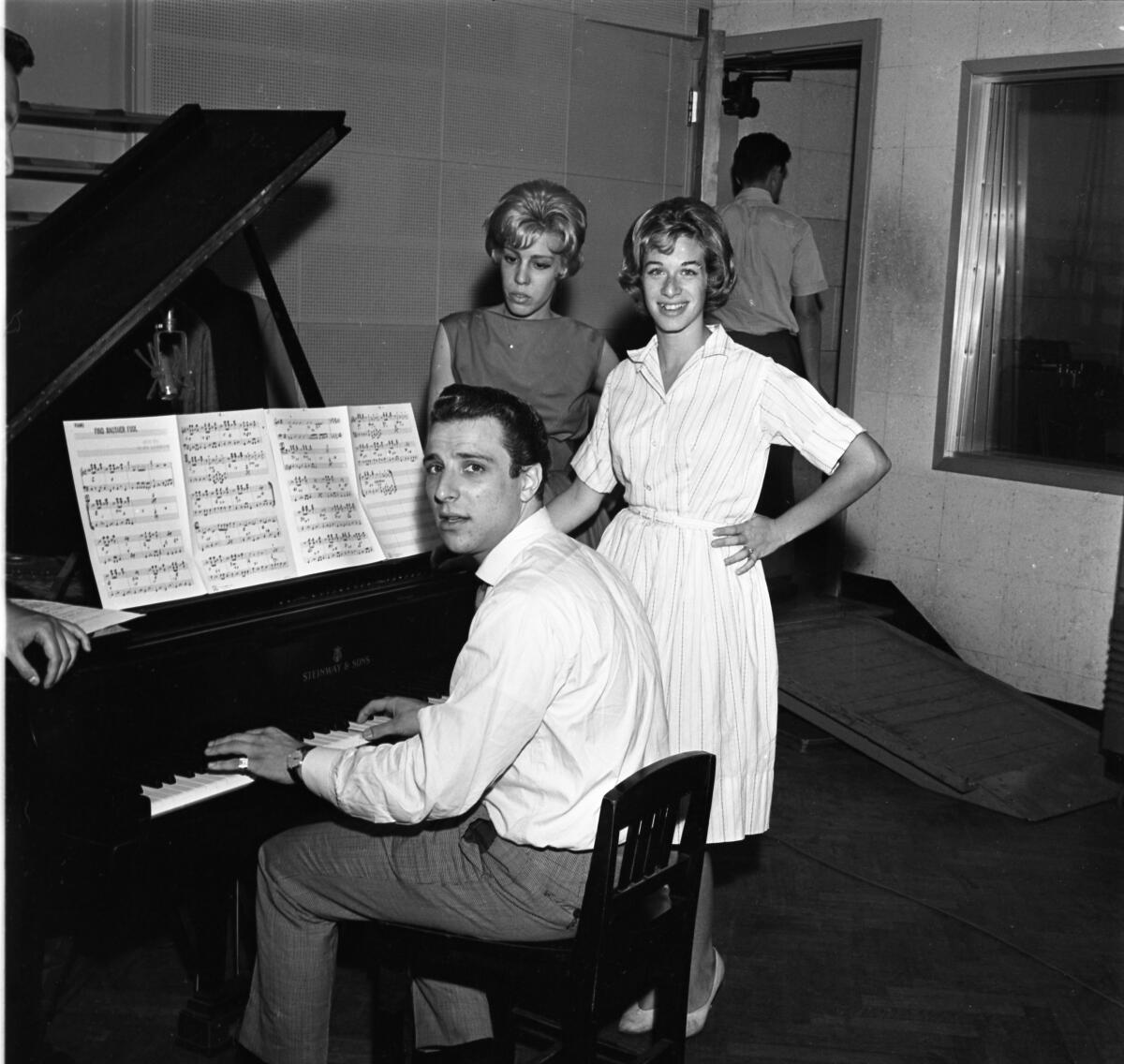 Two women stand next to a man seated at a piano