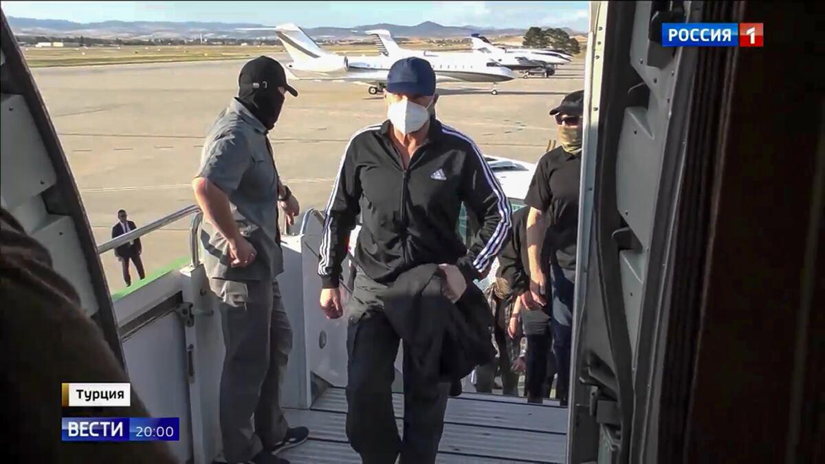 A man in a white mask, dark cap and clothes, walks past another man as he boards a plane, with other people behind him