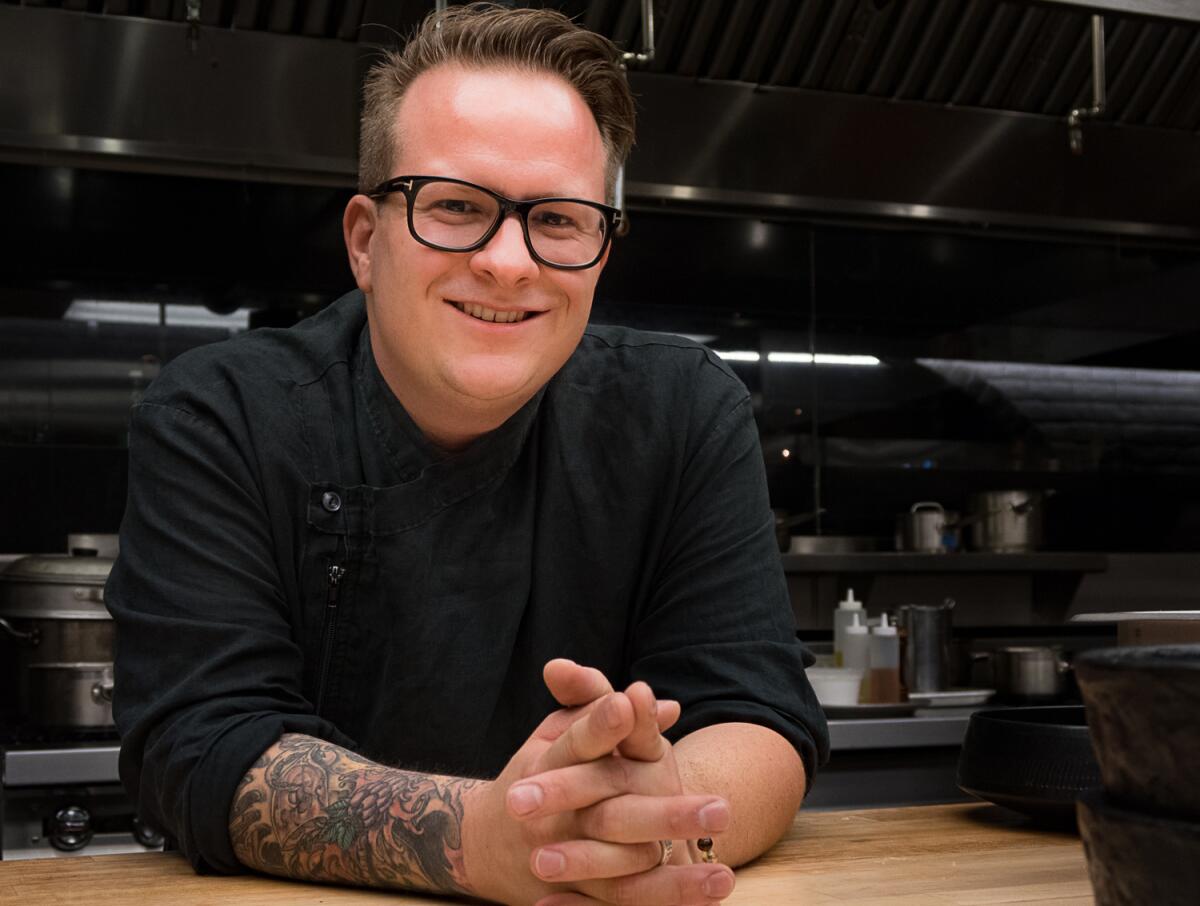 Chef Brian Howard in black chef's jacket and black-rimmed glasses with tattoos visible on his right forearm