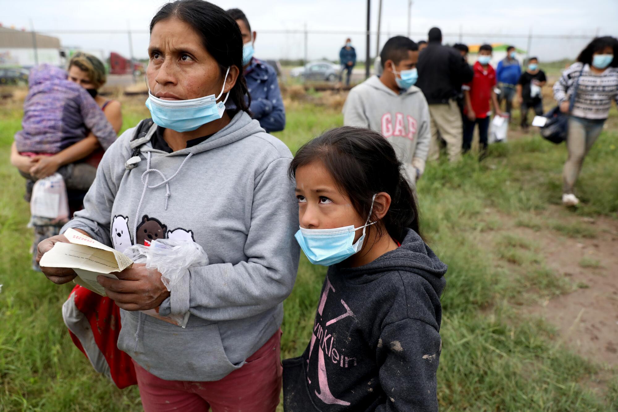 A woman and a girl wait with other people in a grassy area