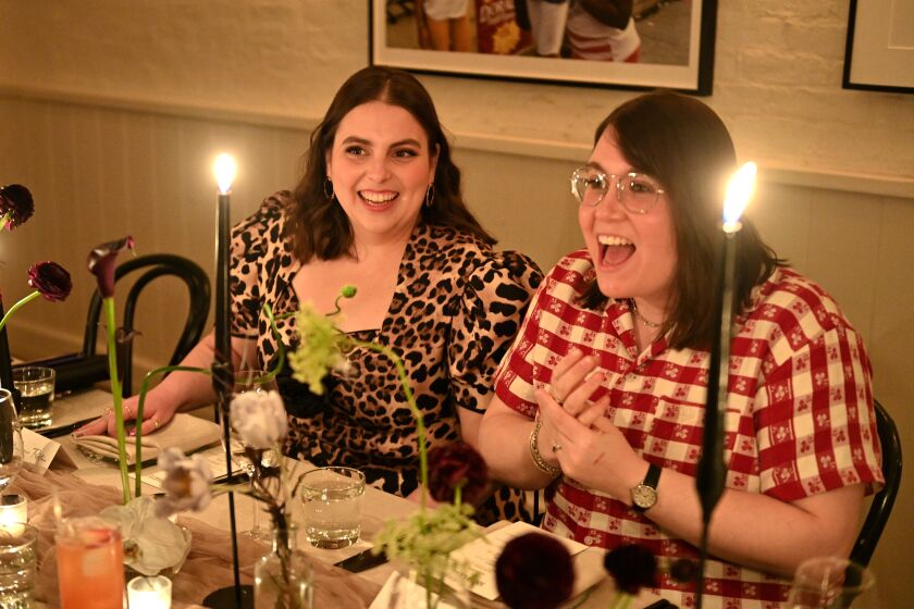 Beanie Feldstein in a leopard-print dress sitting next to Bonnie Chance-Roberts in a red checkered shirt at a candlelit table
