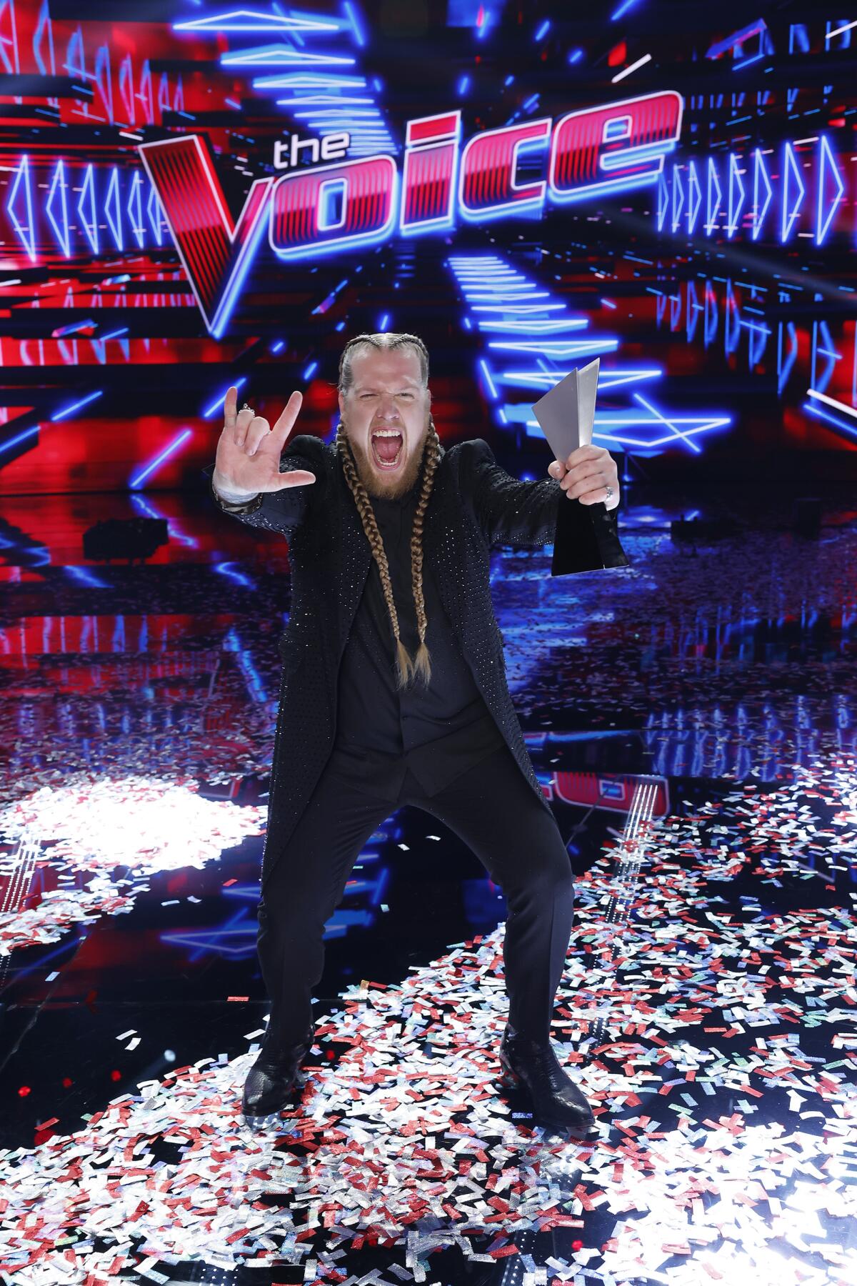 A man with long, blond braids in a black suit holding out a rock sign and a metal trophy standing over confetti