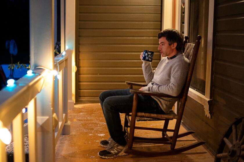 A man sits on a porch in a rocking chair at night, drinking from a mug