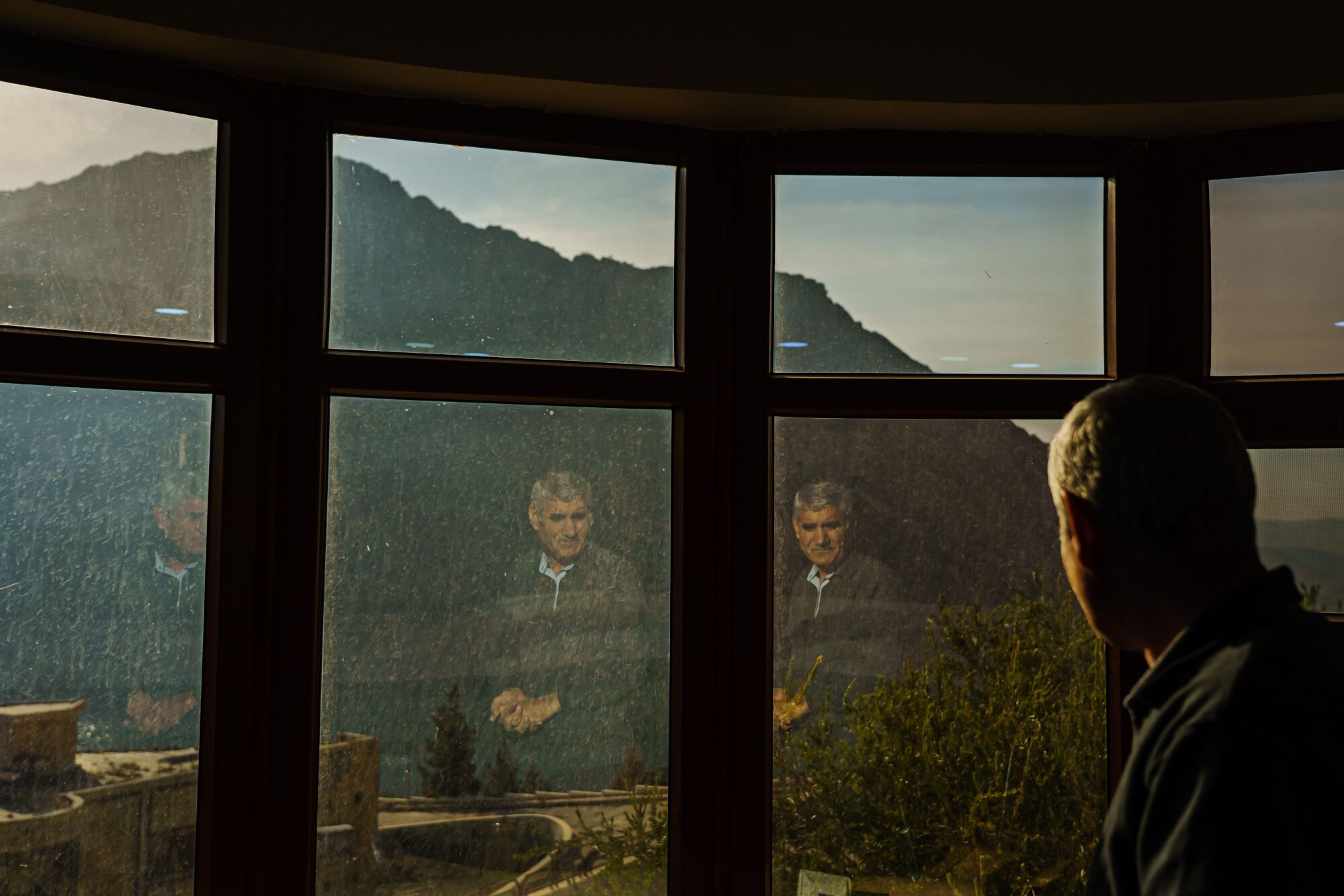 A man looking out onto the dam outside his office is reflected in the glass of his window 