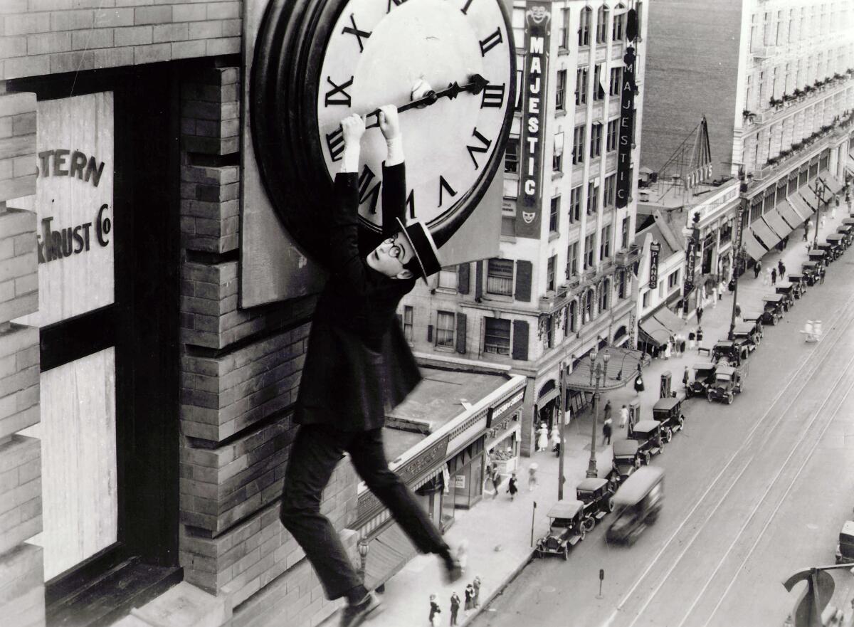 A black-and-white film still in which Harold Lloyd dangles from a large clock grasping one of its arms with his hands. 