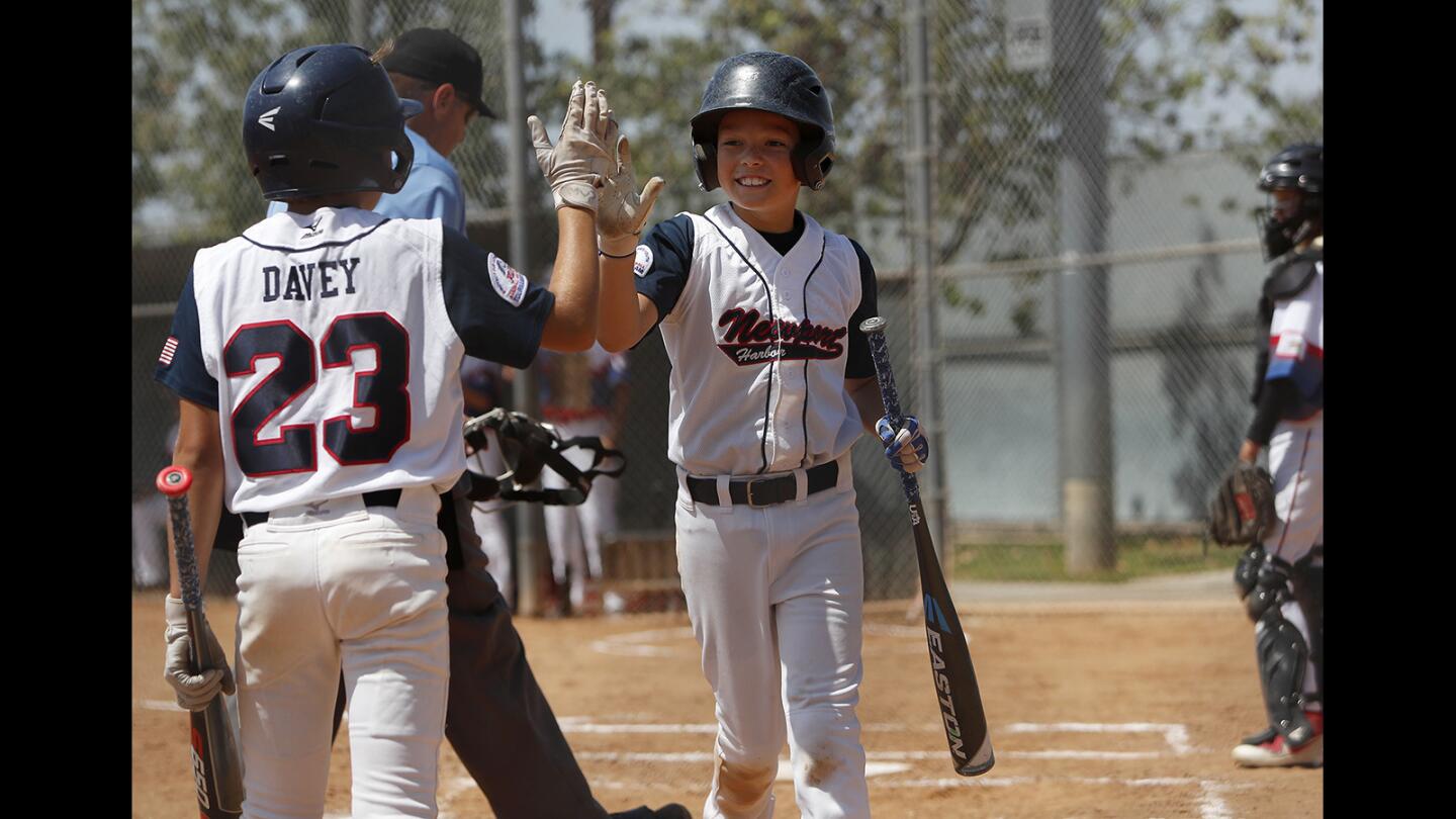 Newport Harbor Baseball Assn.'s Nicolas Montgomery, center, is congratulated by teammate Jack Davey after scoring a run against Dana Point during the fifth inning in the PONY Bronco 12-and-under District tournament at Placentia Champions Sports Complex on Saturday, June 30.