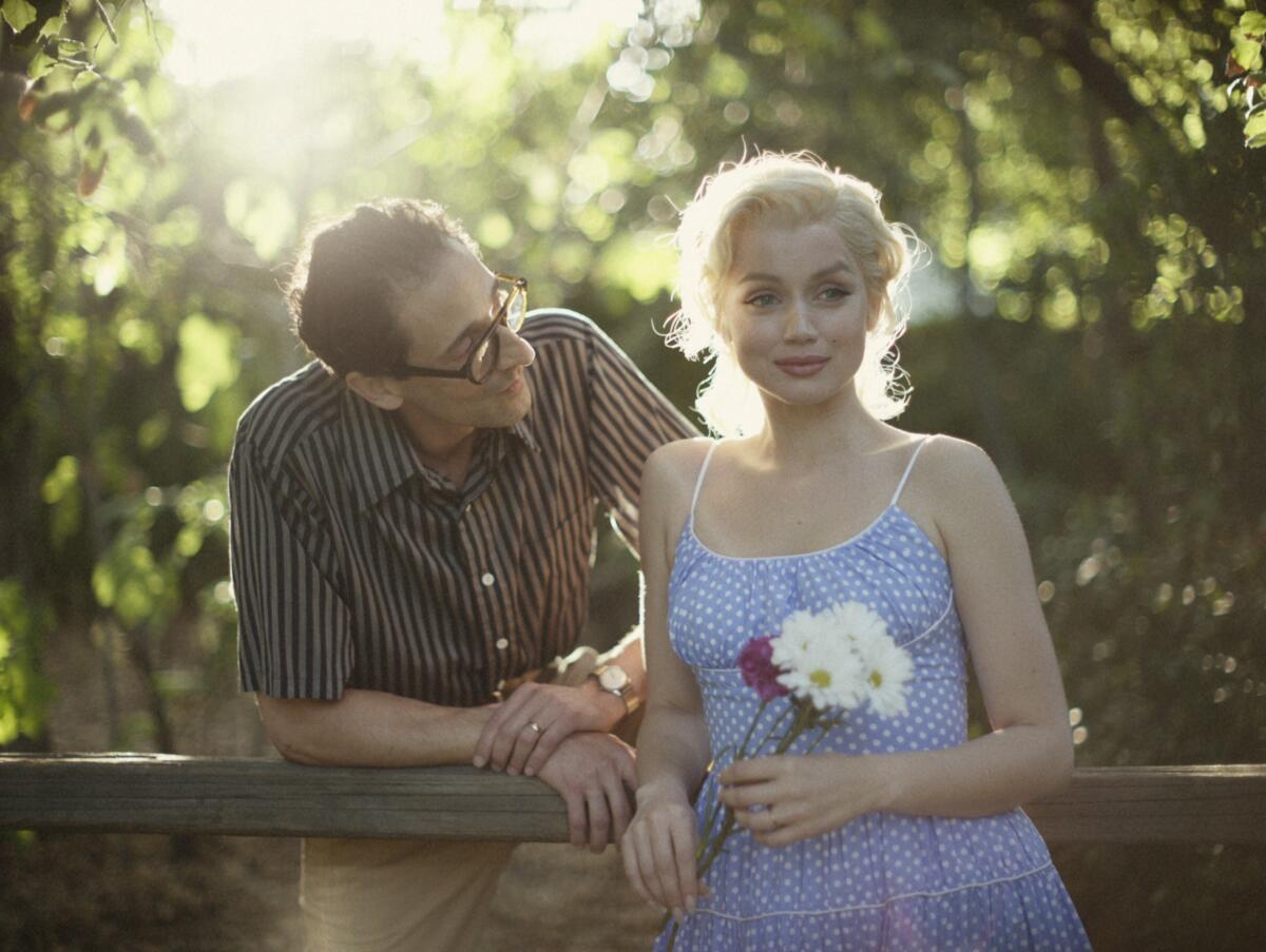 A man in glasses looks at a blond woman wearing a blue dress and holding flowers in the movie "Blonde."
