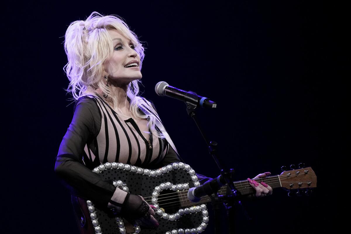 Dolly Parton in a black and beige outfit stands holding a guitar with pearl detailing and singing on a dark stage