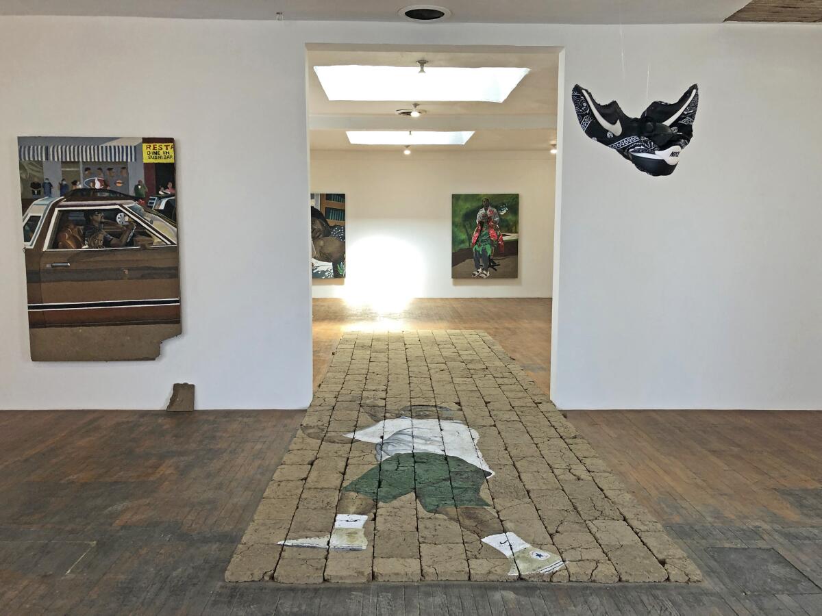 A wide view of a gallery shows paintings on a wall and broad adobe sculpture on the floor that shows a figure of a man