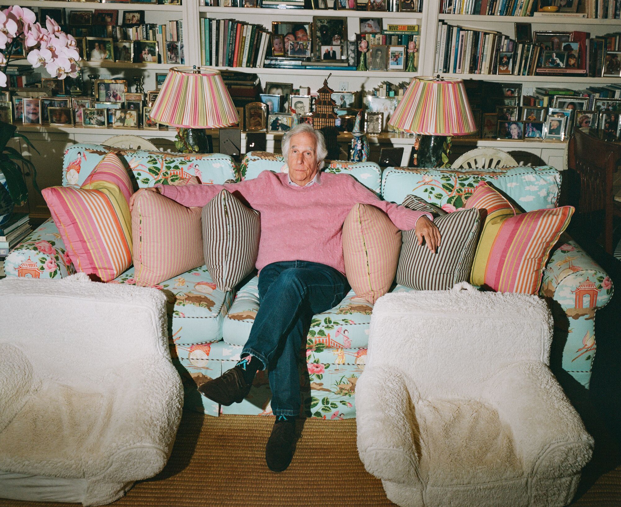 A man in a pink sweater sits on a blue floral couch with a lamp and fluffy white chairs on either side of him