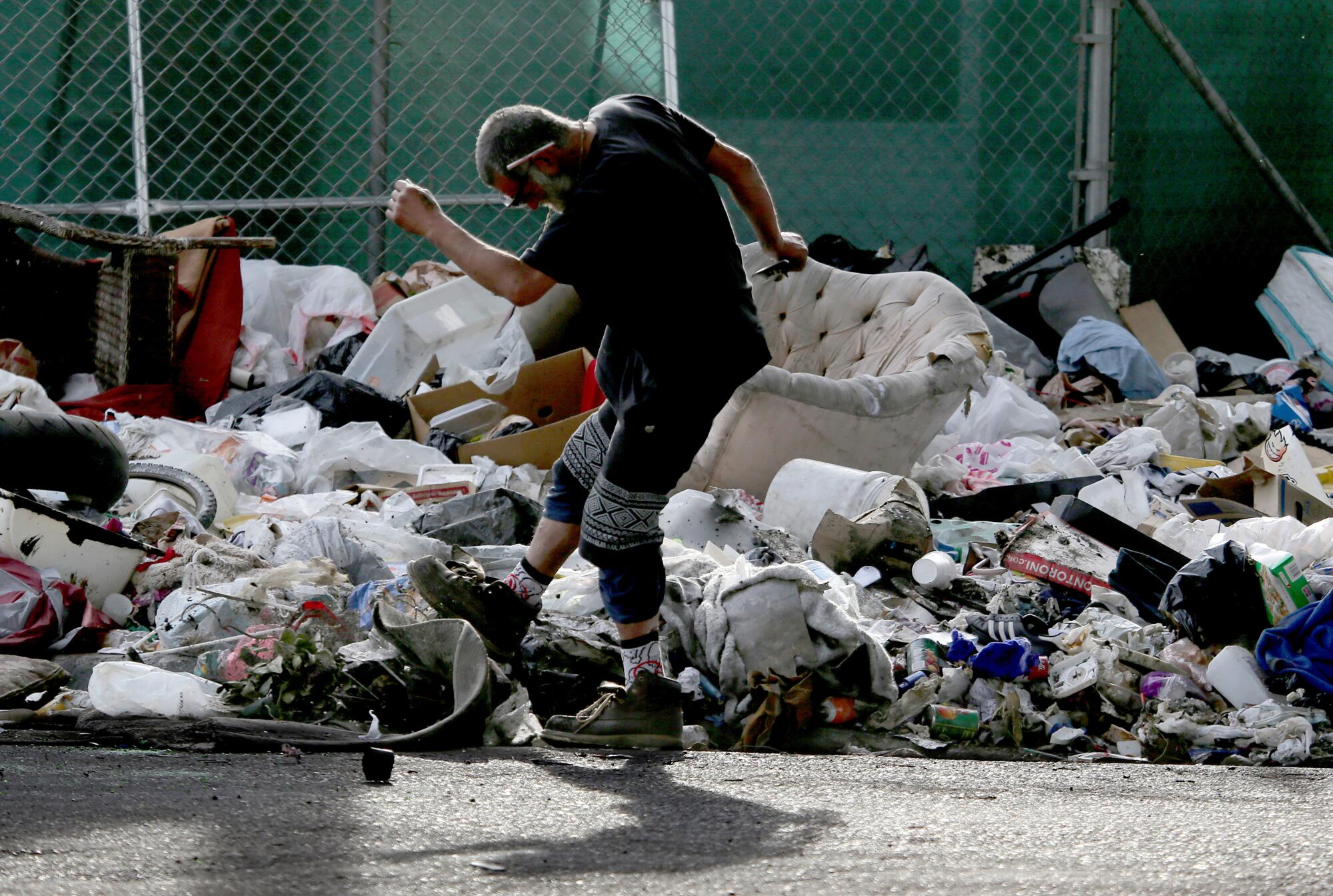 A man lifts his arms and kicks up a foot near a ripped-up couch and other trash piled against a chain-link fence.
