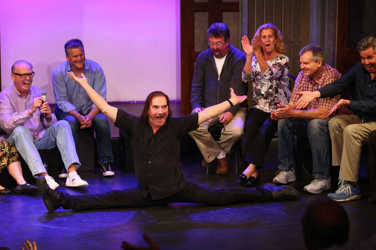 A man sits in a split on a stage surrounded by laughing people