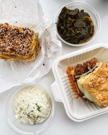 An overhead photo of two biscuit sandwiches with sides of collard greens and grits at Auntie Beulah’s Biscuits & Chicken.