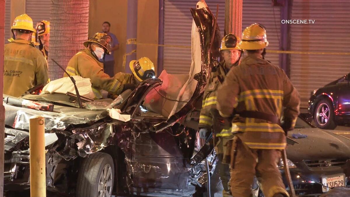 Firefighters stand near a crushed black sedan. 